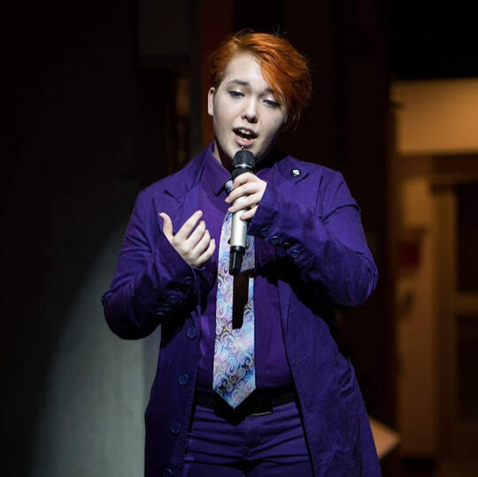 A photo of Bard Nolan at an event, singing for an audience obscured in shadow in the foreground. Photo by James McDaniel